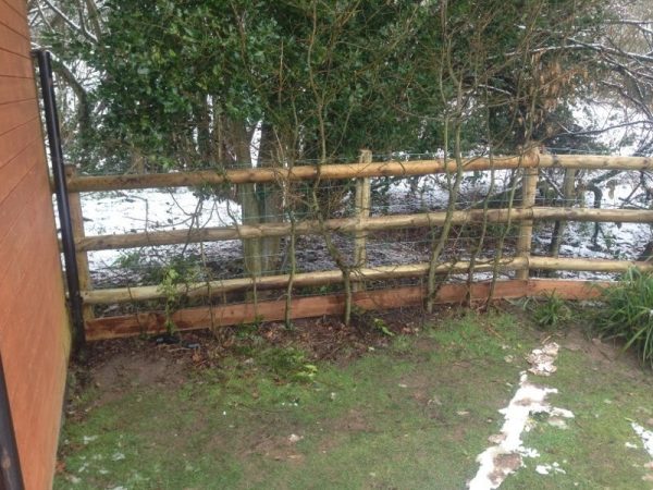 A fence runs along the side of a house. The fence has round fence posts and half round fence posts.