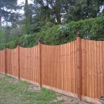 A wooden fence runs along the perimeter of a building. It is a warm wood and stands horizontally by two trees.