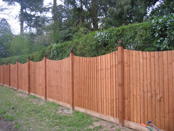 A wooden fence runs along the perimeter of a building. It is a warm wood and stands horizontally by two trees.