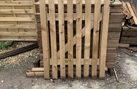 A wooden paling gate is stood horizontally against wooden posts in a stocked supply yard.